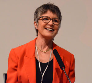 Photograph of Dr Meredith Doig, smiling, wearing red jacket, in front of microphone.